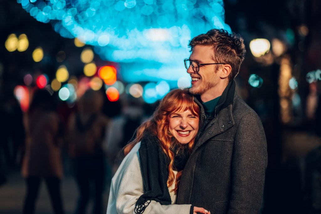 An affectionate couple is standing on city street illuminated by lights and decorations, hugging and laughing