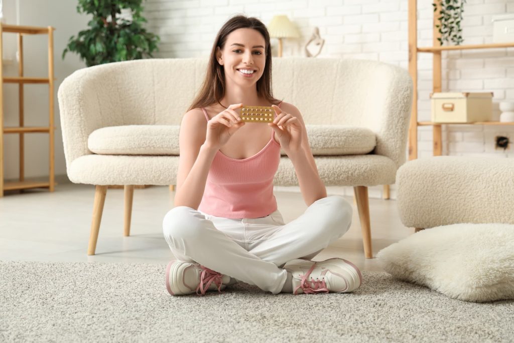 Beautiful young woman with birth control pills sitting on carpet in living room