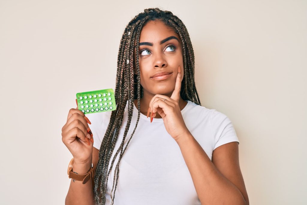 Young african american woman with braids holding birth control pills serious face thinking about question with hand on chin