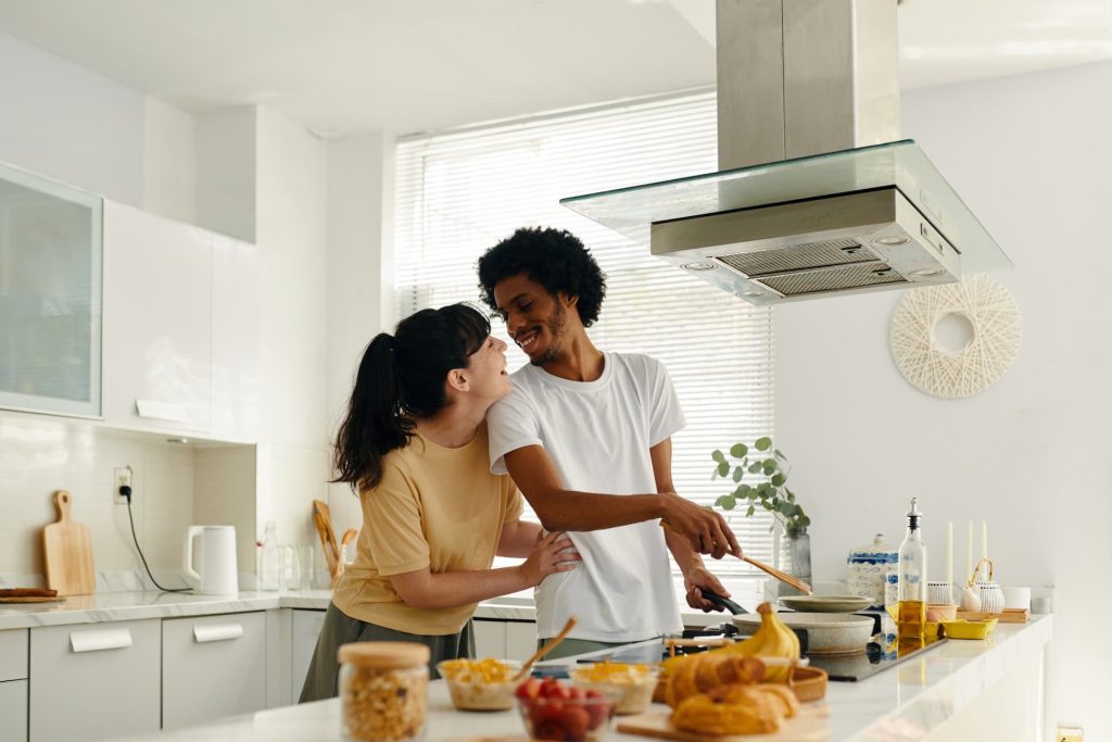 Young cheerful man looking at his wife while standing by electric stove in the kitchen and preparing something tasty for breakfast