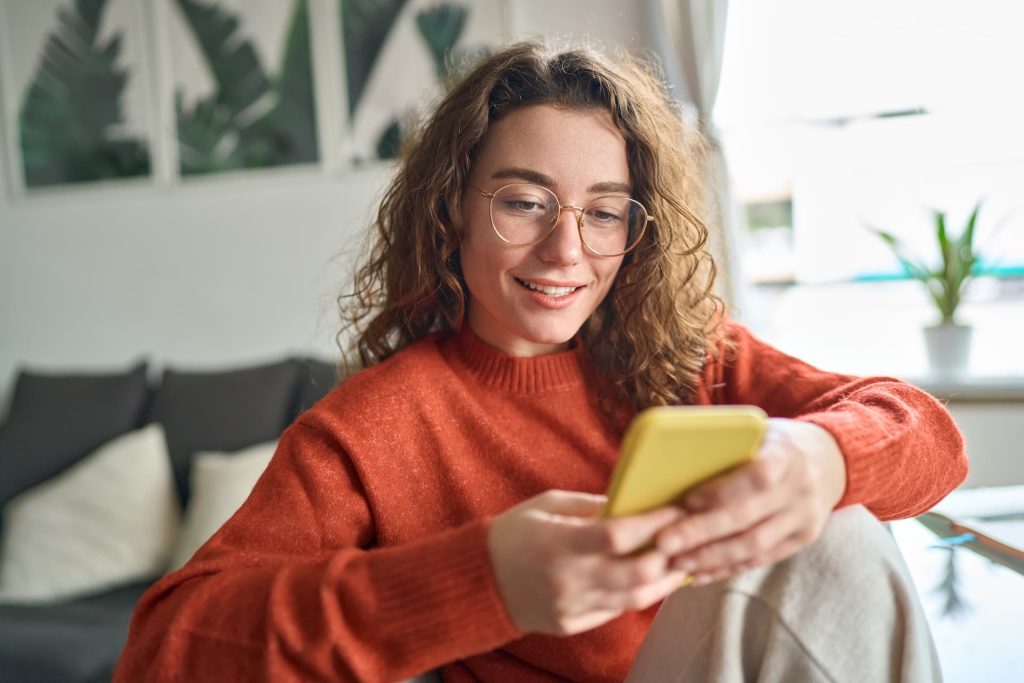 Young smiling woman wearing glasses holding smartphone using cellphone to check pregnancy calculator sitting at home